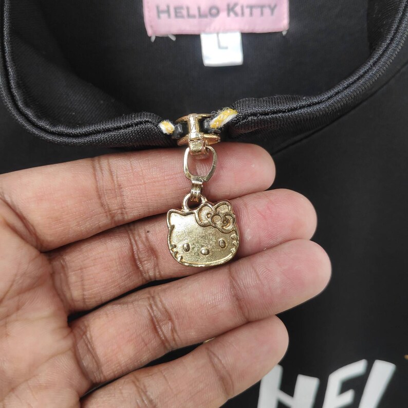 May include: Black zipper pull with a gold Hello Kitty charm. The charm features Hello Kitty's face with a red bow.