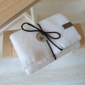 Könnte beinhalten: Eine gefaltete weiße Decke, gebunden mit schwarzem Bindfaden und einem kleinen Anhänger mit einem Herz. Ein kleines braunes Etikett ist an der Decke befestigt. Die Decke liegt auf einer braunen Oberfläche.