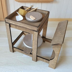 Könnte beinhalten: Miniatur-Töpferscheibe aus Holz mit einem kleinen Tablett mit Werkzeugen und einem Schwamm. Das Rad hat eine silberfarbene Drehfläche und einen passenden Sitz. Das Set ist aus Holz gefertigt und für ein Puppenhaus oder eine Miniatur-Ausstellung konzipiert.