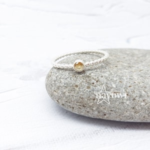 Könnte beinhalten: Ein silberner Ring mit einem perlenbesetzten Band und einem hellgelben Edelstein. Der Ring liegt auf einem grauen Stein, vor weißem Hintergrund. Ein zarter Schmuck.