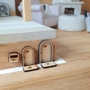 Könnte beinhalten: Miniatur-Holzhausfassade mit einem Briefkasten mit der Aufschrift "POST" und zwei bogenförmigen Türen. Die Türen haben herzförmige Ausschnitte oben und sternförmige Ausschnitte unten. Das Haus ist aus hellbraunem Holz gefertigt.