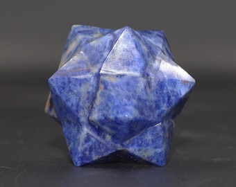 May include: A faceted, blue gemstone sculpture in the shape of a star. The stone has a deep blue color with lighter blue and white veining. The geometric design features sharp, pointed edges and a symmetrical form.