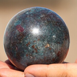 May include: A smooth, dark green and brown sphere with red flecks. The sphere is made of polished stone and has a high gloss finish.