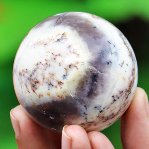 May include: A white and purple agate sphere with brown and black inclusions. The sphere is smooth and polished.