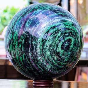 Puede incluir: Una esfera verde, negra y rosa hecha de piedra pulida. La esfera está sentada sobre un soporte de madera.