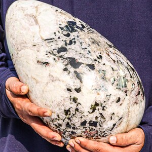 May include: A large, white, oval-shaped stone with black and green flecks. The stone is being held by two hands.