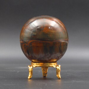 Puede incluir: Una esfera pulida de piedra de ojo de tigre multicolor sobre un soporte de metal dorado con tres patas.