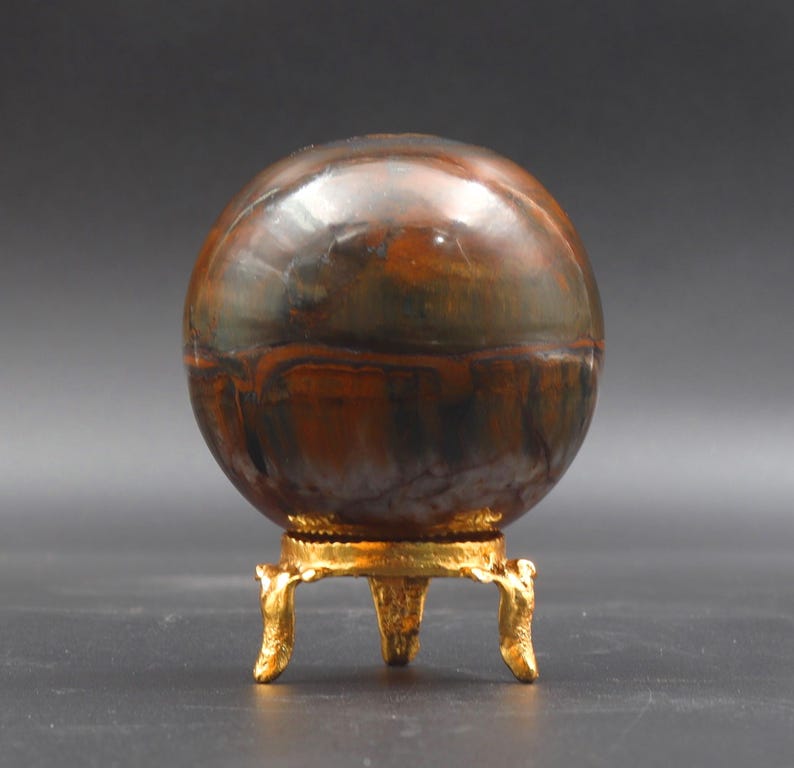 Puede incluir: Una esfera pulida de piedra de ojo de tigre multicolor sobre un soporte de metal dorado con tres patas.