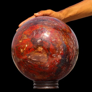 Op de afbeelding: Een grote, gepolijste stenen bol met een complex, gemarmerd patroon van dieprode, gebrand oranje en donkerbruine tinten met doorschijnende kristallijne insluitingen, rustend op een kleine, ronde houten standaard voor presentatie.