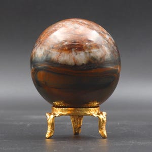 Puede incluir: Una esfera de ojo de tigre pulida, de color marr&oacute;n y blanco, sobre un soporte de metal dorado con tres patas.