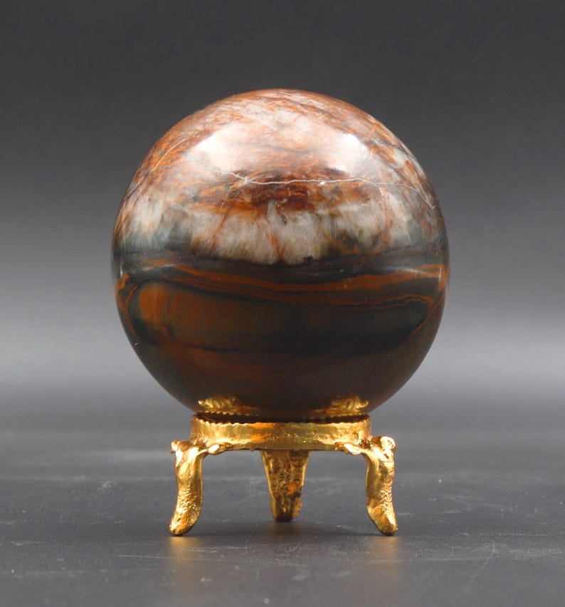 Puede incluir: Una esfera de ojo de tigre pulida, de color marr&oacute;n y blanco, sobre un soporte de metal dorado con tres patas.