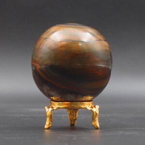 Puede incluir: Una esfera pulida multicolor con rayas marrones, doradas y negras se asienta sobre un soporte de metal dorado con tres patas.