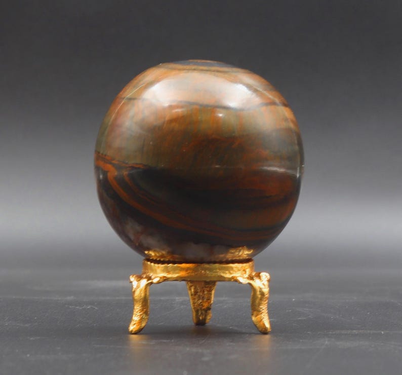 Puede incluir: Una esfera pulida multicolor con rayas marrones, doradas y negras se asienta sobre un soporte de metal dorado con tres patas.