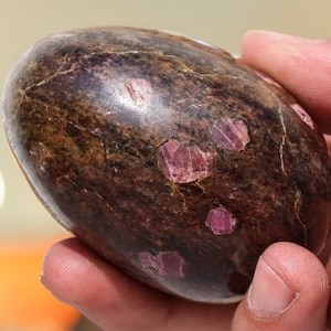 Puede incluir: Una piedra de color marrón oscuro y negro con forma de huevo con cristales rosados incrustados en la superficie.
