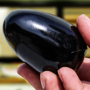 Puede incluir: Un huevo de obsidiana negro liso, pulido a alto brillo. El huevo se sostiene en una mano, mostrando su tamaño y forma.