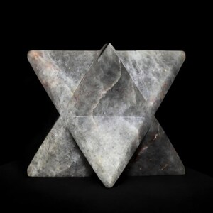 Könnte beinhalten: Eine graue Steinskulptur in Form eines sechsspitzigen Sterns, vor schwarzem Hintergrund. Die geometrische Form hat eine strukturierte Oberfläche mit subtilen Farbvariationen, einschließlich Andeutungen von Braun und Weiß.