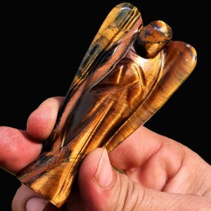Peut inclure: Une figurine d'ange en &oelig;il de tigre sculpt&eacute;. L'ange a des ailes d&eacute;ploy&eacute;es et une expression sereine. La pierre pr&eacute;sente des bandes de brun dor&eacute;, de noir et de jaune, avec une finition polie. La figurine est tenue dans une main sur fond noir.