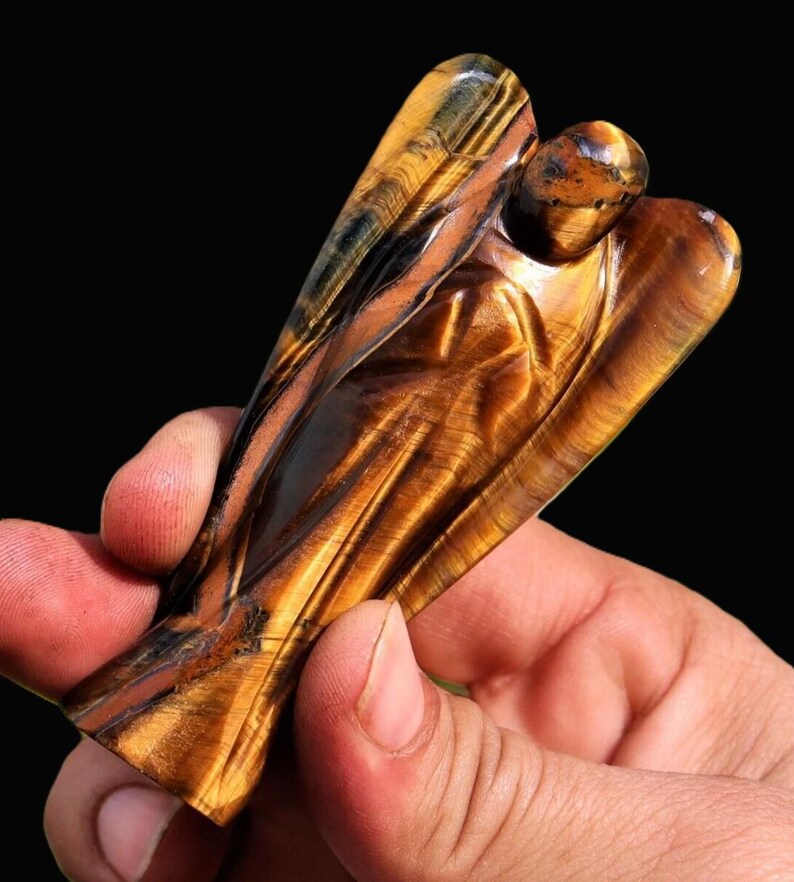 Peut inclure: Une figurine d'ange en &oelig;il de tigre sculpt&eacute;. L'ange a des ailes d&eacute;ploy&eacute;es et une expression sereine. La pierre pr&eacute;sente des bandes de brun dor&eacute;, de noir et de jaune, avec une finition polie. La figurine est tenue dans une main sur fond noir.