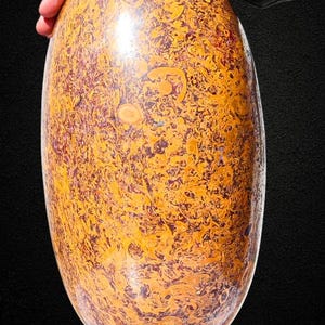 May include: A large, oval decorative stone with a polished surface. The stone has a complex pattern of orange, yellow, and brown swirls and markings. It is mounted on a red wooden base, and a hand is resting on top.