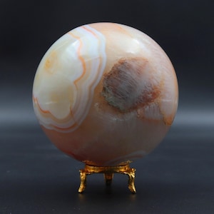 May include: A large, polished agate egg with white and orange banding. The egg is displayed on a gold metal stand with three legs.
