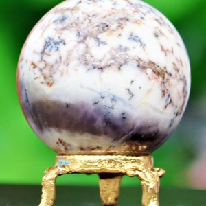 May include: A white and purple agate sphere on a gold metal stand with three legs. The sphere has a swirling pattern of white, purple, and brown.