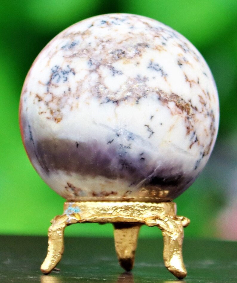 May include: A white and purple agate sphere on a gold metal stand with three legs. The sphere has a swirling pattern of white, purple, and brown.
