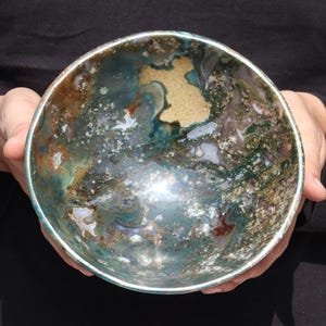 May include: A large, round bowl made of polished green, brown, and white agate. The bowl has a natural, organic pattern and a smooth, shiny surface.