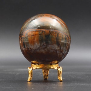 Puede incluir: Una esfera de ojo de tigre pulida sobre un soporte de metal dorado. La esfera tiene rayas marrones, doradas y negras.
