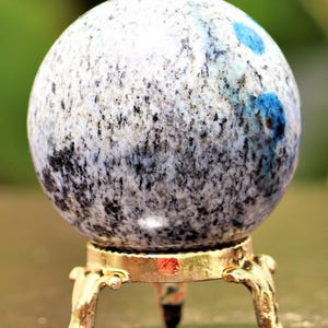 May include: A polished blue and white sphere with black flecks sits on a gold metal stand with three legs. The sphere is a decorative object.