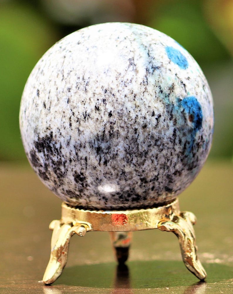 May include: A polished blue and white sphere with black flecks sits on a gold metal stand with three legs. The sphere is a decorative object.