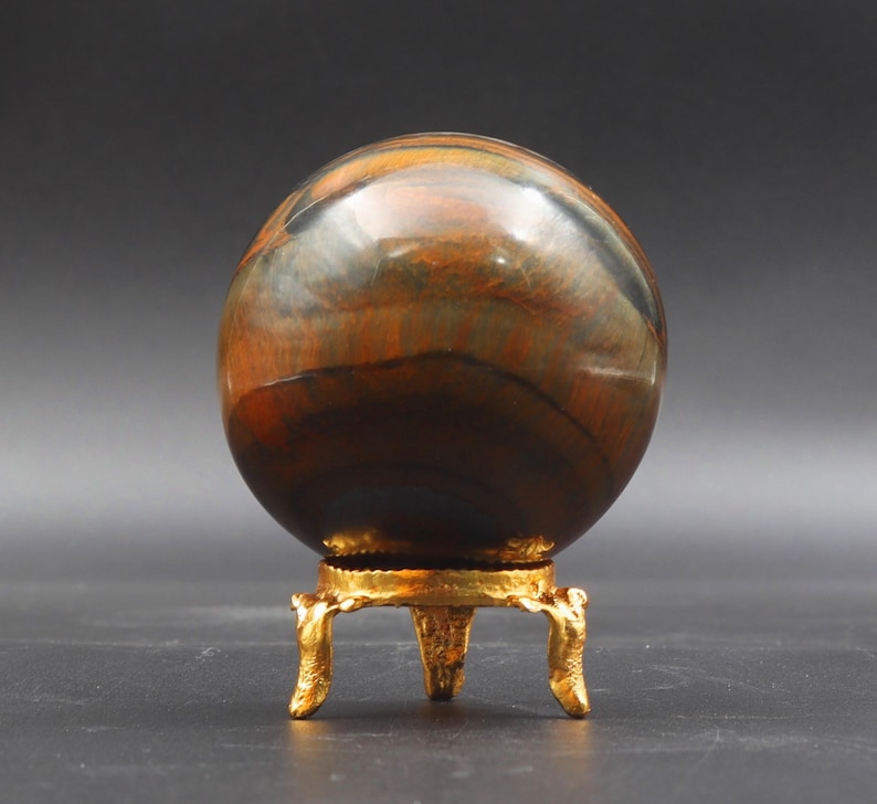 Puede incluir: Una esfera de ojo de tigre pulida, multicolor, sobre un soporte de metal dorado con tres patas. La esfera tiene rayas marrones, doradas y negras.