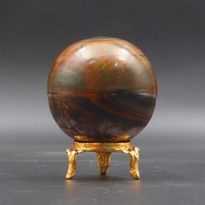 Puede incluir: Una esfera de ojo de tigre pulida sobre un soporte de metal dorado con tres patas. La esfera tiene un patr&oacute;n de color marr&oacute;n, dorado y negro.