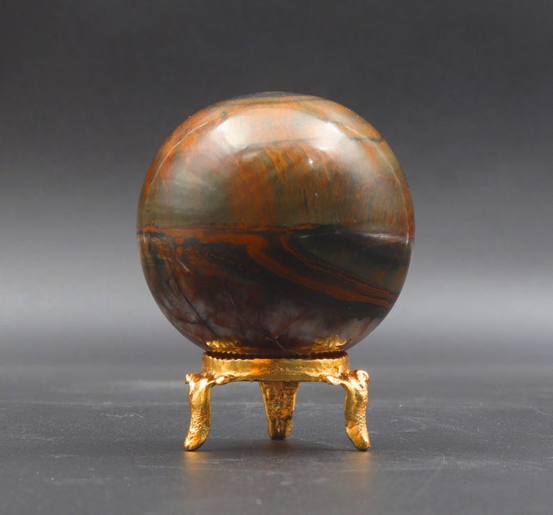 Puede incluir: Una esfera de ojo de tigre pulida sobre un soporte de metal dorado con tres patas. La esfera tiene un patr&oacute;n de color marr&oacute;n, dorado y negro.