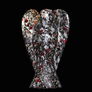 Könnte beinhalten: Eine Steinengelfigur mit ausgebreiteten Flügeln. Die Skulptur besteht aus gesprenkeltem Granit in Schwarz-, Weiß- und Grautönen mit roten Akzenten. Der Engel steht mit gefalteten Händen vor der Brust. Der Hintergrund ist schwarz.