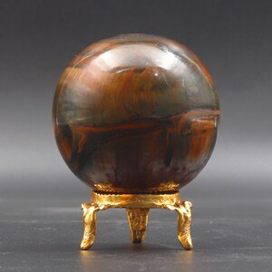 Puede incluir: Una esfera de ojo de tigre pulida, multicolor, sobre un soporte de metal dorado con tres patas.