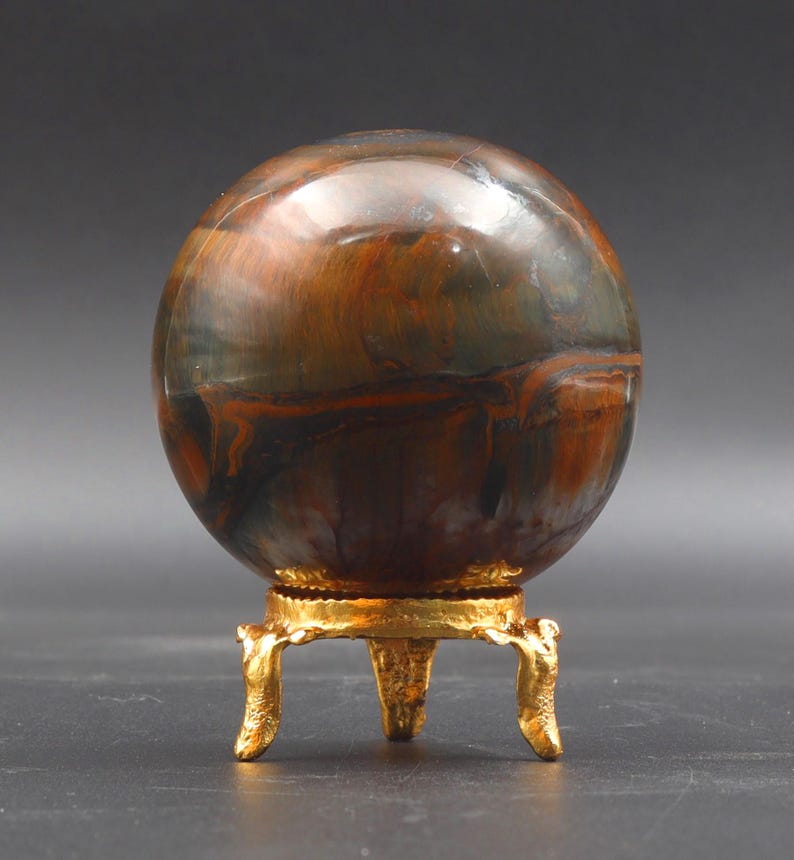 Puede incluir: Una esfera de ojo de tigre pulida, multicolor, sobre un soporte de metal dorado con tres patas.
