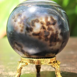 May include: A polished agate sphere with brown and white patterns, displayed on a gold metal stand with three legs.