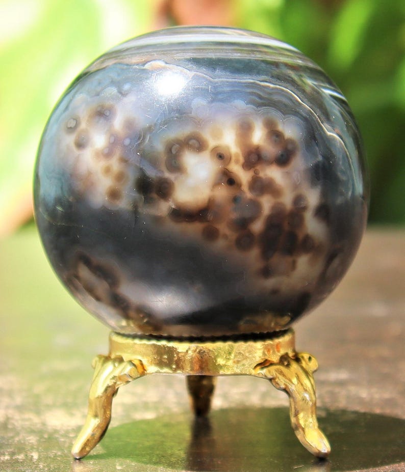 May include: A polished agate sphere with brown and white patterns, displayed on a gold metal stand with three legs.