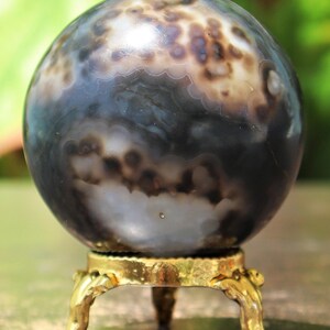 May include: A polished agate sphere with a swirling pattern of black, white, and gray. The sphere is displayed on a gold metal stand with three legs.