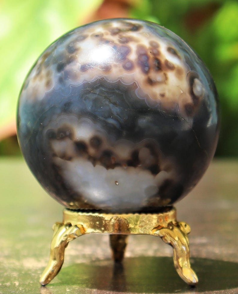 May include: A polished agate sphere with a swirling pattern of black, white, and gray. The sphere is displayed on a gold metal stand with three legs.