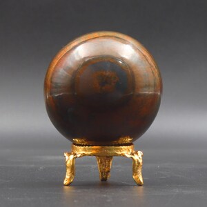 Puede incluir: Una gran esfera pulida de color marr&oacute;n y dorado con matices iridiscentes azules y verdes se asienta sobre un soporte de metal dorado con tres patas.