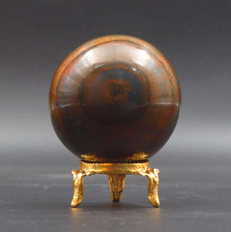 Puede incluir: Una gran esfera pulida de color marr&oacute;n y dorado con matices iridiscentes azules y verdes se asienta sobre un soporte de metal dorado con tres patas.