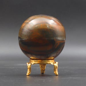 Puede incluir: Una esfera de ojo de tigre pulida, de color marr&oacute;n y negro, sobre un soporte de metal dorado con tres patas.