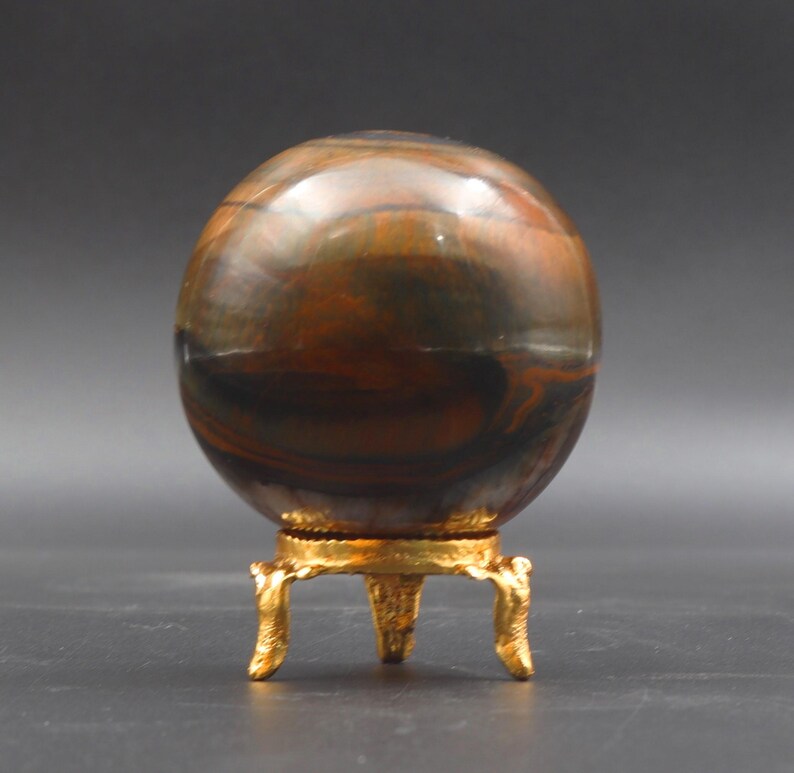 Puede incluir: Una esfera de ojo de tigre pulida, de color marr&oacute;n y negro, sobre un soporte de metal dorado con tres patas.