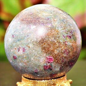 May include: A spherical gemstone with a mottled appearance, featuring a mix of green, brown, and reddish-pink hues. The polished surface of the sphere is mounted on a gold-coloured base, showcasing the natural variations in the stone.