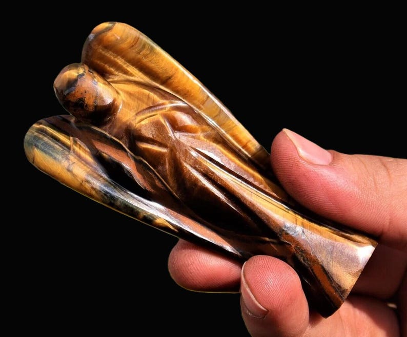 Peut inclure: Une figurine d'ange sculpt&eacute;e en pierre d'&oelig;il de tigre. L'ange a des ailes d&eacute;taill&eacute;es et une expression sereine. La pierre pr&eacute;sente des bandes de marron, d'or et de noir.