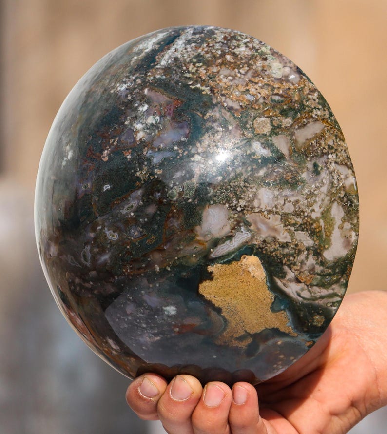 May include: A large, polished sphere of green, brown, and white moss agate. The sphere has a smooth, shiny surface and intricate patterns of colour.