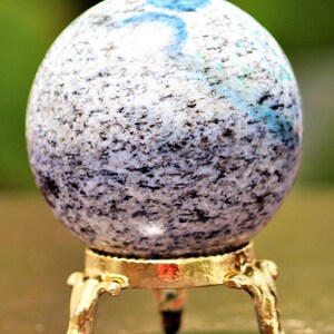 May include: A white and gray speckled sphere with blue and green flecks sits on a gold metal stand with three legs.