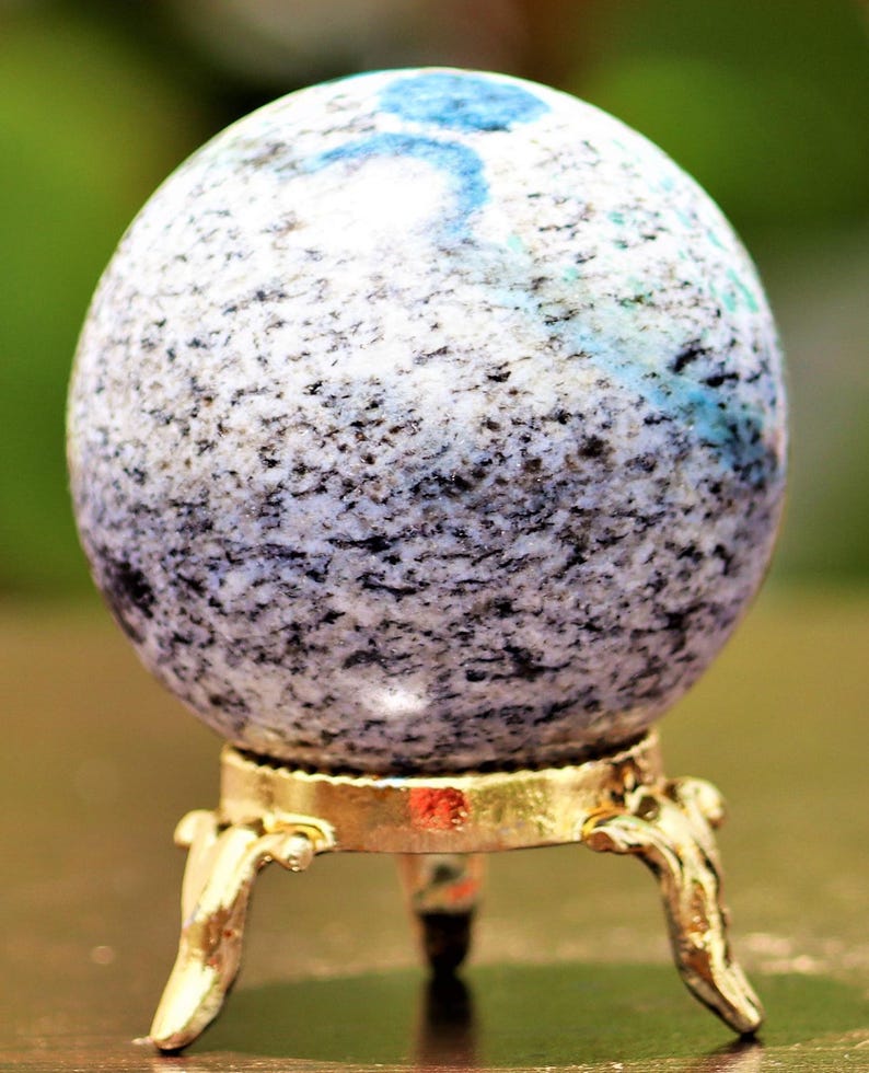 May include: A white and gray speckled sphere with blue and green flecks sits on a gold metal stand with three legs.
