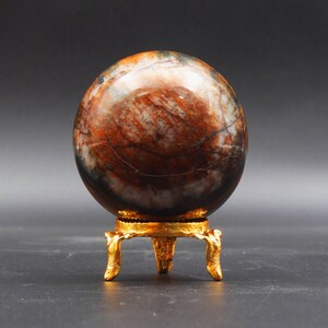 Puede incluir: Una esfera pulida de &aacute;gata multicolor sobre un soporte de metal dorado con tres patas. La esfera tiene un patr&oacute;n de m&aacute;rmol marr&oacute;n, blanco y negro.